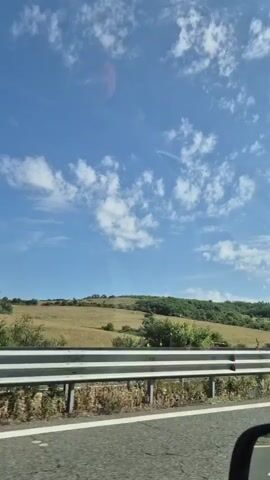 The video shows a roadside view from a moving vehicle. A guardrail separates the road from a grassy embankment. Beyond the embankment, a rolling green hill is visible under a blue sky with scattered white clouds. The sun appears to be bright. There are no people or other vehicles visible in the frame. The video is shot at a slight angle, suggesting the vehicle is moving.