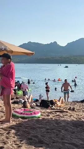 A beach scene with people swimming in the water and relaxing on the sand. The water is calm, and the sky is clear with mountains in the background. Some people are playing with inflatable toys in the water, while others are sunbathing or sitting on the sand. A boat is visible in the water with several people on it. The overall atmosphere is one of leisure and recreation.
