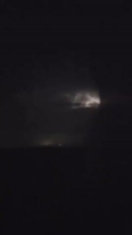 The video depicts a dark, stormy night over the ocean. Lightning illuminates the clouds in the distance, casting a faint glow on the water. A distant light, possibly from a ship or a coastal town, is visible on the horizon. The waves are dark and indistinct. The overall atmosphere is ominous and dramatic.