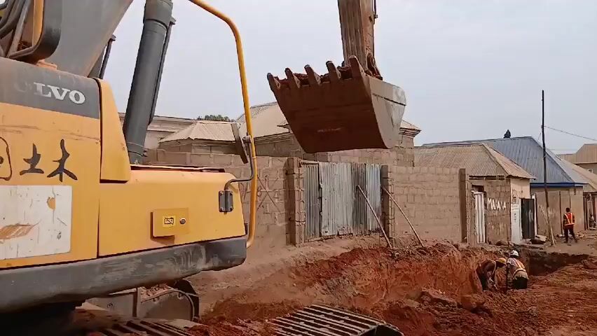 An excavator is digging a trench along a road with brick buildings in the background. Two construction workers are in the trench. There are other people standing on the road, and a white truck is parked nearby. The camera moves to show the tracks of the excavator. Then the camera moves to show the side of the truck and a person in jeans and black shoes walking by. The camera then gets very close to the person's fingers.