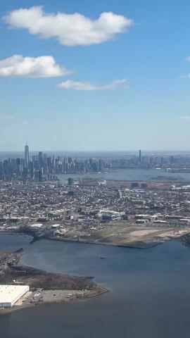 The video shows an aerial view of a city skyline with the Freedom Tower prominently visible. The sky is blue with scattered white clouds.  Below, there is a mix of urban development with a river or large body of water running through it. Various industrial buildings and infrastructure can be seen, including bridges and open areas. The camera appears to be moving, giving a panoramic view of the cityscape.