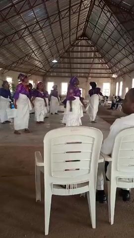 In a spacious room with a high, corrugated metal roof, a group of women are dancing. They are uniformly dressed in white wraps and purple tops, with matching head coverings. The room has multiple windows providing natural light. In the foreground, two white plastic chairs are visible, with a person seated in one, facing towards the dancers. Other individuals are seated further back, observing the dance. The floor appears to be a light brown dirt.