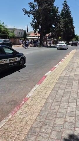 A police car with lights on is parked on the side of a street. Several people are standing on the sidewalk across the street from the police car, some of whom appear to be looking at something on the road. Two cars are parked further down the street, and several other people are visible walking and standing near them. The weather is clear and sunny.