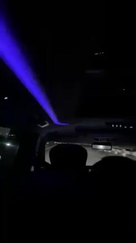 The video shows the interior of a vehicle at night. The camera is positioned in the back seat, looking forward towards the front of the vehicle. A blue light strip is visible along the ceiling of the car. The rear-view mirror and the back of the front seats are also visible. Through the windshield, a dark road with other vehicles and streetlights can be seen. The car appears to be in motion. The overall lighting is dim, mostly lit by the blue light and the headlights of other vehicles.