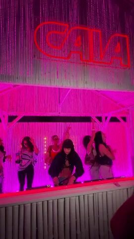 A video captures an energetic scene in a pink-lit indoor venue. A large red neon sign spelling "CAIA" is visible against a wall of light-colored vertical slats. Below the sign, a group of seven women are positioned behind a white, slatted counter that extends across the frame. The woman in the center, wearing a dark long-sleeved top, leans forward over equipment, appearing to be a DJ. To her left and right, five other women are actively dancing, raising their arms, and moving to the music. They are dressed in various casual outfits, including light tops, dark pants, and some wearing sunglasses. On the far right, partially visible in the foreground, is what appears to be a person's arm and shoulder, suggesting the video is shot from a crowd perspective. The entire area is bathed in a vibrant pink light.
