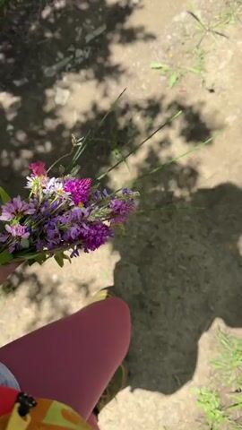 A person is walking outdoors in what appears to be a park or wooded area. They are holding a bouquet of wildflowers in their left hand. The bouquet consists of various purple and pink flowers, along with some greenery. The person is wearing bright pink leggings and a green and yellow patterned shoe. The ground is a mix of dirt and grass, with dappled sunlight filtering through trees. A shadow of the person and a tree are visible on the ground.