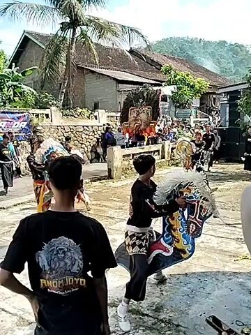 The video shows a group of people participating in what appears to be a traditional Indonesian dance or performance outdoors. Several performers are dressed in colorful costumes, some holding large, decorative fans. They are dancing in a paved area in front of a building. There is a small crowd watching the performance, including a person wearing a black shirt with a graphic on the back who appears to be observing the dance. In the background, there is a banner, trees, and residential buildings, indicating the event is taking place in a village or small town setting. The camera moves from the dance to people gathered near a building with green pillars, showing the audience is comprised of a mix of people, including women in hijabs.