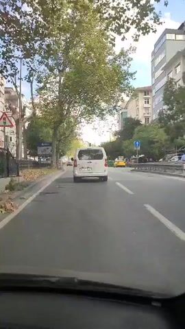 A white van is driving down a city street. The van is in the center of the frame. There are trees lining the street on both sides. There are buildings on either side of the street. The sky is blue with some clouds. There are other cars on the road, including a yellow taxi. The video appears to be shot from inside another vehicle.