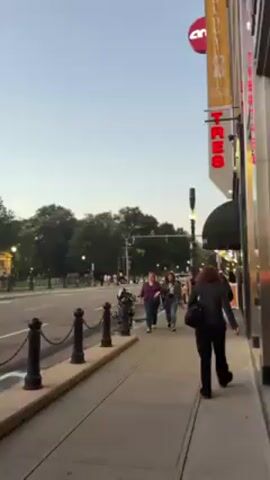 The video shows a city sidewalk beside a multi-lane street with a park visible across the street. There are several pedestrians walking along the sidewalk. A sign with the word "TRES" in red neon letters is visible on a building. The sky is light. There are black bollards connected by chains along the edge of the sidewalk. A motorcycle is parked near the bollards.