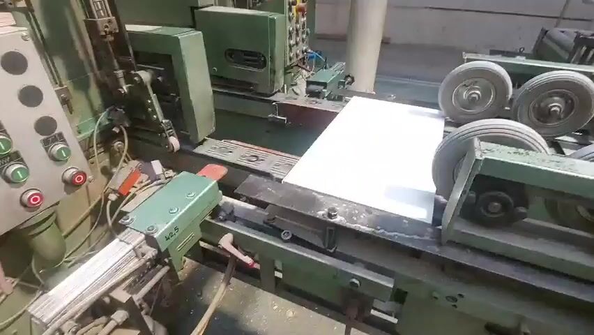 A white panel is being processed by industrial machinery in a workshop. The panel is situated on a conveyor belt-like system that moves it through the machine. The machinery is predominantly green, with various mechanical components visible, including rollers, cutting blades, and control panels with buttons. The environment appears to be an indoor industrial setting, possibly a factory or workshop, with visible dust or debris on the machinery, suggesting active use. The camera is positioned to capture the processing of the panel from a slightly elevated angle. There are no people visible in the frame.