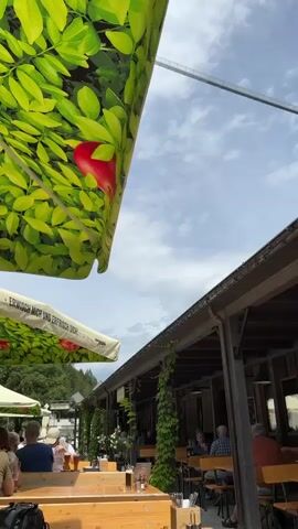 This video shows an outdoor scene with tables and chairs set up in what appears to be a restaurant or beer garden. People are seated at the tables, some with drinks and food. A large umbrella with a green and red apple pattern is visible in the foreground, providing shade. In the background, a long, narrow pedestrian suspension bridge stretches across the sky. The sky is cloudy with patches of blue. Green vines are growing on the side of a building in the middle ground.