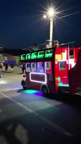 A small, red, double-decker bus-shaped amusement ride is moving slowly at night on a paved surface, possibly a street or pathway, illuminated by streetlights and the ride's own neon lights. Several people are walking alongside and behind the ride. In the foreground, a person wearing a gray t-shirt and shorts walks away from the camera, holding hands with a child in a yellow t-shirt and dark shorts. Behind them, several other people are visible walking in the same direction. The ride itself has green, red, and yellow lights. The environment appears to be an outdoor area with some buildings and fences visible in the background, suggesting a park, fairground, or similar entertainment venue. The sky is dark, indicating it is nighttime.