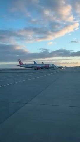 The video shows a Jet2 airplane at an airport, stationary on a runway or taxiway. The plane is silver with red and white Jet2 branding. There are several ground vehicles near the plane. The sky above is a mix of blue and clouds, with the scene illuminated by what appears to be either sunrise or sunset. The camera appears to be moving forward and slightly to the right along marked lines on the pavement.