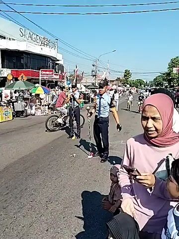 The video depicts an outdoor scene on a sunny day, likely a street transformed for a public event. In the foreground on the right, a woman in a pink hijab and light brown top is seated or crouching, holding a mobile phone and observing the activity ahead. A child is partially visible beside her. In the middle of the road, a marching band is present, with members dressed in dark blue uniforms featuring light blue or white sashes. Several band members are visible, some carrying or adjusting large drums, including bass drums with a red swirl emblem. Crowds of people line both sides of the road, with bicycles parked along the left curb. Colorful market stalls with canopies are visible further down the right side of the road. Buildings and utility poles are present in the background under a clear blue sky.