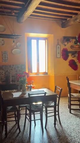 The video shows the interior of a rustic-looking room, possibly a dining area or kitchen, with a focus on its decor and design. The room features exposed wooden beams on the ceiling, and the walls are painted in warm, earthy tones, possibly textured to give them an aged appearance. The floor is tiled with small, irregular patterned tiles. Multiple wooden tables and chairs are arranged in the room. The first table has a marble top, adorned with a vase of red flowers. Kitchenware like copper pots and pans, and decorative plates are hanging on the wall.  A window provides a view of an exterior stone building. The overall impression is that of a cozy, old-world Italian style kitchen or dining room.
