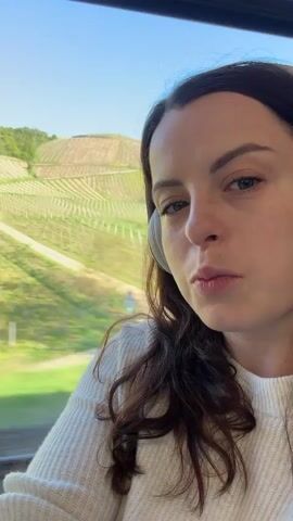 A woman is seen in close-up, likely seated on a train or other moving vehicle, looking towards the camera. She has fair skin, dark brown hair, and is wearing a white knit sweater and white headphones. Outside the window, a hillside covered in vineyards scrolls by, blurred by the motion. The sky is visible, appearing blue with some clouds. Part of a window frame and a colorful graffiti-covered object are also visible in the foreground, slightly out of focus. The woman maintains a neutral or slightly pensive expression.