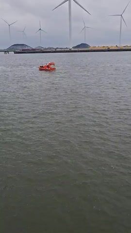 A wide shot shows a waterway on an overcast day. Several wind turbines stand in the distance beyond a flat, green land mass. An orange life raft with two people in it drifts slowly across the water. To the left, a gray, metal, tower-like structure is visible on a gray platform above the water. The water is dark and has small ripples.