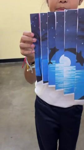 A child in a white t-shirt and navy pants holds an accordion-folded paper picture in front of them. The picture depicts a colorful scene of dolphins, a sunset, and palm trees, transitioning from night to day. The child moves the folded paper, displaying the alternating images. The scene is indoors, with a neutral-toned wall and visible classroom-like furniture. A child-sized bracelet is on the right arm.