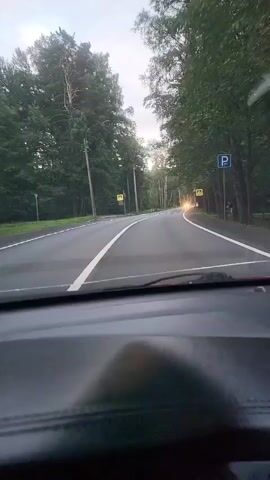 The video is shot from inside a car, looking forward through the windshield. The car is driving on a paved road that curves to the right. The road is lined with trees on both sides. There are white lane markings on the road. On the right side of the road, there are a few white poles and a blue parking sign. On the left side of the road, there are two yellow pedestrian crossing signs. In the distance, the headlights of an oncoming car are visible. The sky appears overcast. The interior of the car is visible in the foreground, including the dashboard. The time stamps indicate the video is a short clip, possibly from a dashcam recording.