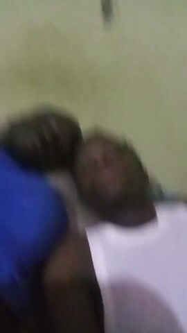 Two dark-skinned men are lying down together, with a pale green wall visible in the background. The man on the left wears a blue shirt, while the man on the right wears a white tank top. Both men are looking directly at the camera with a neutral expression.