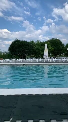 The video shows an outdoor swimming pool on a sunny day with scattered clouds. The pool is filled with clear blue water and has white tiling around the edge. Beyond the pool, there is a row of empty lounge chairs, some white umbrellas, and lush green trees. The scene is peaceful and suggests a recreational area.
