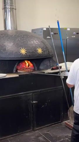 A person wearing a white shirt and black pants is standing in front of a wood-fired pizza oven. The oven is domed and decorated with black tiles and golden sun symbols. Flames are visible inside the oven, cooking a pizza. The person is holding a pizza peel, which they are using to rotate or retrieve the pizza from the oven. To the left of the oven, there is a stainless steel exhaust pipe. To the right, there is a black cabinet with doors. In the background, there is a stainless steel refrigerator with stacked pizza boxes on top. Further to the right, there are more stainless steel appliances. To the left of the oven, there are stacks of pizza boxes, a refrigerator filled with food containers, and several plates of pizza on a counter. The environment appears to be a kitchen or restaurant setting.
