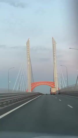 This is a video of a vehicle driving across a large, modern cable-stayed bridge. The bridge has two tall, white pylons connected by numerous white cables to the roadway. An orange, arched structure spans beneath the roadway, illuminated with pinkish-orange lights. The sky is overcast with soft clouds, suggesting either dawn or dusk. The road is paved and marked with white lines. There are guardrails on either side of the road, and streetlights are visible. A white semi-truck is driving ahead of the camera vehicle, and a few other vehicles can be seen in the distance. The overall scene is one of travel and engineering, with the bridge being the dominant feature. The video does not contain any visible text. Based on the provided location context, this bridge is likely in Maloe Okulovo, Russia. The time of day appears to be either early morning or late evening due to the soft, diffused light and the overcast sky.