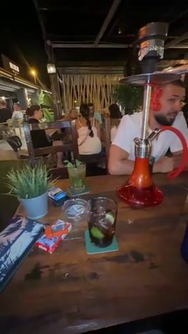 A man and a woman are sitting at a table in an outdoor restaurant. The man is wearing a white shirt and the woman is wearing a white top. A hookah is on the table between them, along with several drinks and cigarettes. The man is looking at the camera and the woman is making a peace sign with her fingers. The background shows other people sitting at tables and a dimly lit ceiling. The environment appears to be a bar or restaurant with a tropical ambiance.