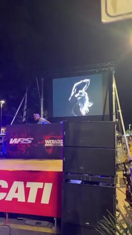 A man stands behind a DJ booth with a laptop and headphones. On the large screen behind him, a motorcycle rider is shown in action, seemingly on a race track, with quick cuts between different angles. The DJ booth is branded with "WRS" and "WDW20". In front of the DJ booth, a red banner with the word "DUCIAII" in large white letters is visible. Two stacks of large black speakers are positioned to the right of the DJ booth. The scene appears to be an outdoor event at night, with stage lighting illuminating the setup.
