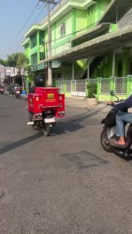 A sunny day with clear skies, a street scene features two motorcycles. In the foreground, a person on a dark scooter rides to the right. Behind them, another person on a white motorcycle with a red delivery box on the back travels forward. The red box has yellow text and logos. Buildings line the street, one of which is a green building with white trim and awnings. Trees and a utility pole are also visible.