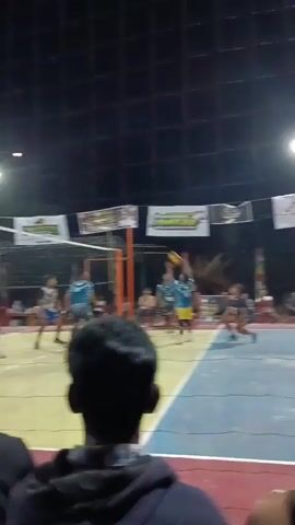 The video shows a volleyball game at night. The court is lit by overhead lights. There are several players on the court, wearing blue and white uniforms. Spectators are visible in the foreground, partially obscuring the view of the game. A volleyball is in the air. The players are in motion, with some attempting to hit or block the ball. Banners are hanging above the court. The overall atmosphere appears to be competitive and energetic.