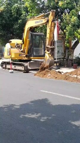 The video shows a yellow Komatsu excavator digging a hole on the side of a road. A red truck is driving past on the road, carrying a load of what appear to be sacks of cement. A person on a motorcycle is also seen riding past the excavator. The scene is set outdoors with trees visible in the background.