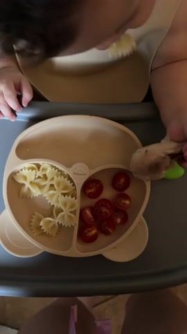 A close-up shot shows a young child in a high chair being fed. The child is wearing a bib and has a segmented plate in front of them, divided into two sections. One section contains a pile of bow-tie pasta, and the other holds several sliced cherry tomatoes. In the child's hands, they are holding a piece of cooked chicken. The child's fingers are interacting with the chicken, and their other hand is resting on the plate. A green silicone food holder is also visible on the tray. The background is blurred, but appears to be an indoor setting. The overall scene depicts a mealtime for a young child.