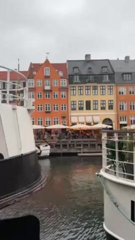 The video shows a waterfront scene in Copenhagen, Denmark on an overcast day. The camera is positioned between two boats, partially obscuring the view of the colorful buildings lining the canal. The buildings are several stories high, with orange, yellow, and red facades and white-framed windows. A restaurant with outdoor seating is visible along the waterfront, with people dining under awnings. The water in the canal is calm. The sky is grey and overcast.