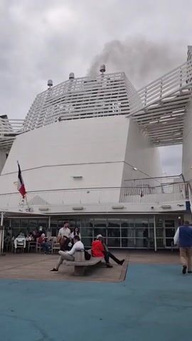 A wide shot shows the aft section of a large white ship or ferry under an overcast sky. Two dark smokestacks on top of the ship's white structure emit dark grey smoke. Below the smokestacks, the ship's upper structure features multiple horizontal white slats. On a deck below, a French flag is visible on the left side. Behind large glass windows on the deck, several people are seated at tables in an interior space. In the foreground, on a blue deck, four men are seated on a light-colored bench. One man in a plaid shirt stands to the left of the bench, holding a large string instrument, possibly a double bass. To the far right, a man in a blue vest walks away from the camera.
