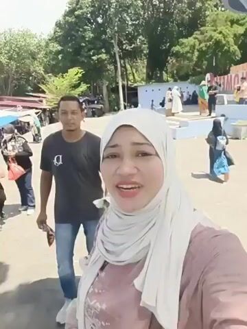 The video shows a family of three posing for a selfie. A woman in a light-colored hijab is in the foreground, smiling at the camera. Behind her, a man wearing a dark t-shirt and jeans waves. To the man's left, a young girl in a hat and blue dress stands with a bottle of water. The family appears to be outdoors in a public area, possibly a park or tourist attraction, with trees and other people visible in the background. There is a large, light-blue structure with red lettering in the background. The lighting suggests it is daytime.
