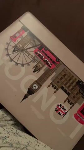 The video begins with a close-up of a light-colored laptop with a London-themed design. The design features iconic London landmarks such as the London Eye, Big Ben, and red double-decker buses. The camera then pans to the laptop's keyboard and trackpad. In the next scene, the laptop screen turns on, displaying a vibrant blue lock screen with a field of lavender under a clear sky. The time is shown as 02:49 on Sunday, 20 September. A hand with grey nail polish is visible touching the trackpad, interacting with the laptop. The environment appears to be indoors, with a soft brown surface and patterned fabric visible in the background.
