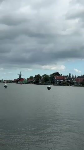 The video shows a wide shot of a calm body of water. The sky is overcast with dramatic clouds. Along the distant shoreline, there is a row of traditional Dutch windmills. Their sails are visible and appear to be stationary. In the water, there are several white buoys. The buildings on the shore are painted in various colors, predominantly green and red, with dark roofs. The overall atmosphere is serene and historic.