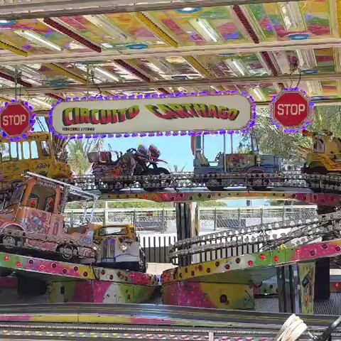 The video shows a colorful carnival ride called "Circuito Carthago." It features various miniature vehicles such as trucks, tractors, and motorcycles on elevated, tilting platforms. A woman wearing a crop top and black pants stands on one of the platforms, seemingly assisting or supervising the ride. The ride's sign hangs above, flanked by stop signs, and the background shows a blue sky and trees. The scene suggests an amusement park or fairground setting.