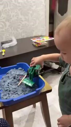 A child is playing with a toy excavator in a blue tray filled with grey sand. The child is wearing a green shirt and is standing next to the tray. The excavator is green and black and has a movable arm and bucket. The tray is sitting on a small wooden table. Behind the tray is a dark brown table with some colorful books on it. The wallpaper has a grey and white floral pattern.