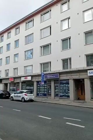 The video captures a street scene in front of a white, multi-story building with many windows. The building has ground-level shops with signage. Several cars are parked along the street, including a white hatchback, a dark sedan, and a red sedan. A small, white utility vehicle is driving down the street away from the camera. There are also streetlights and traffic signals visible. The sky is cloudy.