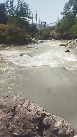 A fast-moving river flows through a rocky, natural landscape. The water is murky and churns as it encounters obstacles in its path, creating small rapids. The riverbanks are covered with vegetation and large rocks. In the background, a power line tower and other industrial structures are visible, suggesting the presence of human development near the natural environment. The sky is clear and the lighting indicates a daytime scene.
