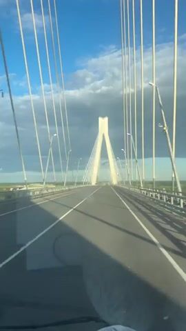 This media file captures a view from a vehicle driving across a modern cable-stayed bridge. The primary subject is the bridge itself, with its striking white pylons and numerous cables stretching across the frame, creating a sense of immense scale and engineering prowess. The road surface is asphalt, marked with white lane lines, indicating it's an active transportation route. The surrounding landscape is visible to the sides and in the distance, featuring lush green trees and a wide body of water, likely a river, given the location context of Im. Voykova, Russia.

The time of day appears to be late afternoon or early evening, suggested by the quality of light and the long shadows cast by the bridge's structures and the vehicle. The sky is a vibrant blue, interspersed with dramatic, greyish-white clouds, hinting at a dynamic weather system. The overall atmosphere is one of movement and journey, with the camera seemingly in motion as it traverses the bridge.

Notable details include the presence of streetlights along the bridge and a hint of industrial elements or buildings in the far distance on the horizon. The scene evokes a feeling of awe at the scale of human construction against the natural backdrop. There is no visible text within the media file. The activity is simply driving across the bridge, offering a perspective of travel and the structure's grandeur. The image implies a sense of open space and a transition across a significant geographical feature.