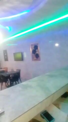 The video shows the interior of a restaurant. The restaurant has tables and chairs with orange, black, and wooden hues. There is a television on the wall. The lighting is colorful, with blue and green neon lights. The walls are decorated with framed artwork and are textured. The video is taken from behind a counter. On the counter are a phone and a menu with a QR code.