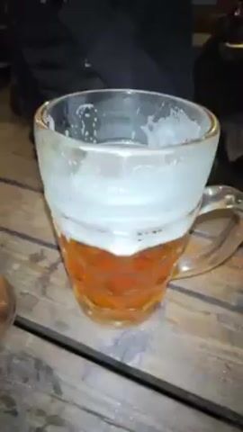 The video shows a wooden table with two large mugs of beer on it. The mugs are filled with amber liquid and topped with white foam. A person's hands move around the mugs, briefly obscuring the view. Another person, wearing a black jacket, sits at the table smoking a cigarette. The setting appears to be a pub or outdoor seating area. There are other people in the background.