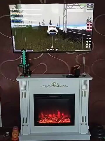 The media file shows a room with a television screen displaying a video game, likely Roblox, featuring a white car driving on a road with trees and buildings in the background. Below the TV is a white electric fireplace with a realistic flame effect. A green toy sword, resembling one from Minecraft, stands on top of the fireplace mantel. The room has brown wallpaper with a swirling pattern. The scene appears to be indoors, suggesting it's likely daytime or evening, as artificial light is prominent. There are no people visible in the frame. The overall impression is one of a cozy, decorated living space, possibly a child's room, set up for entertainment. The visible text on the TV screen is part of the game interface, displaying chat messages and game statistics.
