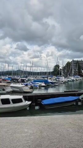 The video shows a marina filled with numerous boats docked on a cloudy day. In the foreground, a white boat with a green canopy is tied to a wooden dock. To the right of this boat, another boat is covered with a blue tarp. Further out in the marina, a dense collection of sailboats with tall masts is visible, along with various other motorboats, many of which are covered. In the background, lush green hills or mountains rise under a dramatic, overcast sky. The water appears calm, reflecting the cloudy sky and the boats. The overall impression is one of a quiet, perhaps pre-storm, atmosphere in a scenic waterfront location.