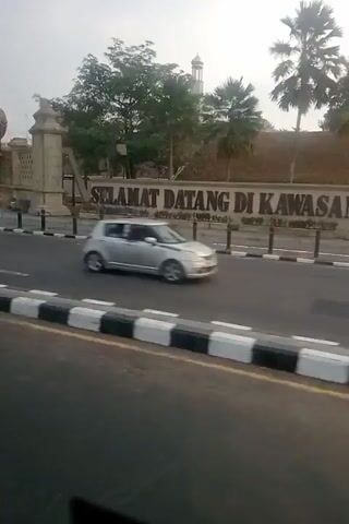 This video shows a street scene with traffic. In the foreground, there is a white car driving on the right side of the road. A traffic light is visible on the left side of the frame. In the background, there is a large sign that reads "SELAMAT DATANG DI" which means "WELCOME TO" in Malay. There are trees and buildings on either side of the street. Several motorcycles and other cars are also visible on the road. The sky is overcast.