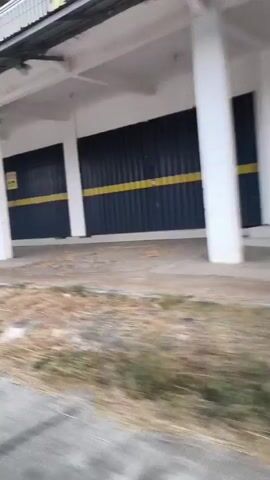 The video shows a side view of a building with a blue and yellow facade, possibly a row of shops, with dark metal shutters. There is a yellow sign visible on one of the shutters. The building appears to be situated alongside a road, with dry grass and some vegetation in the foreground. The camera is moving, suggesting it is capturing footage from a vehicle. The environment seems to be somewhat rural or undeveloped, with trees and brush visible beyond the building. The lighting suggests it may be daytime, possibly overcast or late in the day.