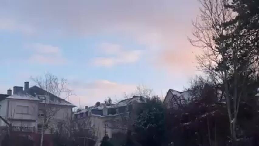 The video shows a wintery landscape with buildings and trees covered in snow. The sky is a mix of light blue and pink hues, suggesting either sunrise or sunset. The camera pans slowly to the right, revealing more trees and rooftops. There are no people visible in the video. The overall scene is peaceful and quiet.